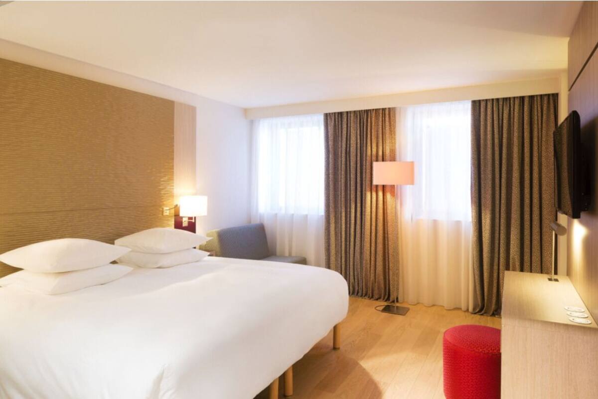 A hotel room with a bed, a desk, a chair, a TV, and a window with curtains.