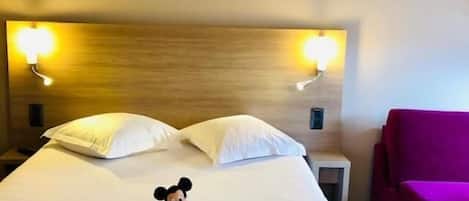 A hotel room with a bed, pillows, a Mickey Mouse plush toy, a nightstand, a lamp, and a purple chair.