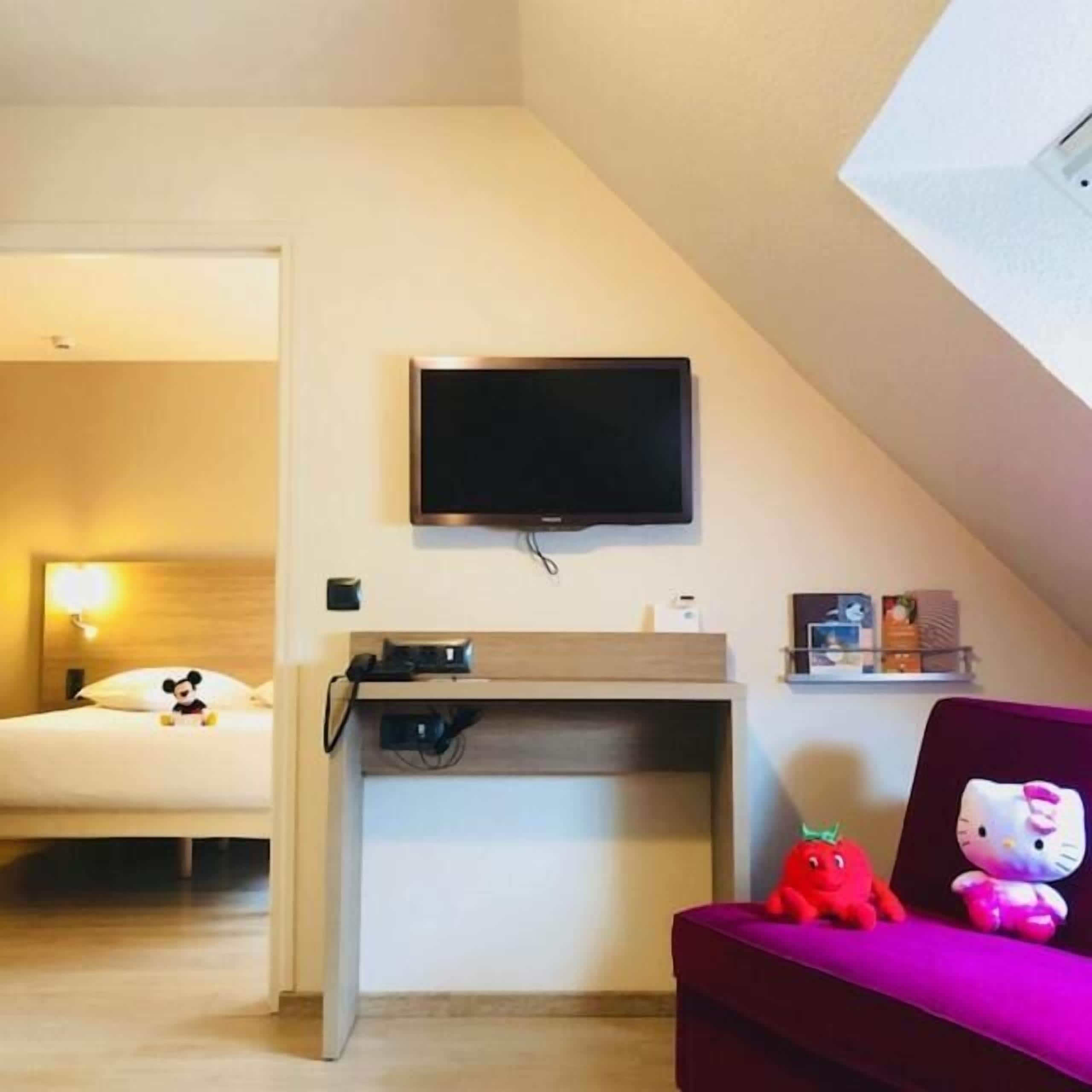 A hotel room with a bed, a desk with a flat-screen TV, a small shelf with items, and a red plush toy on a purple cushion.