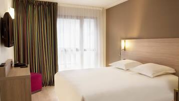 A hotel room with a bed, a desk with a telephone, and a window with curtains.