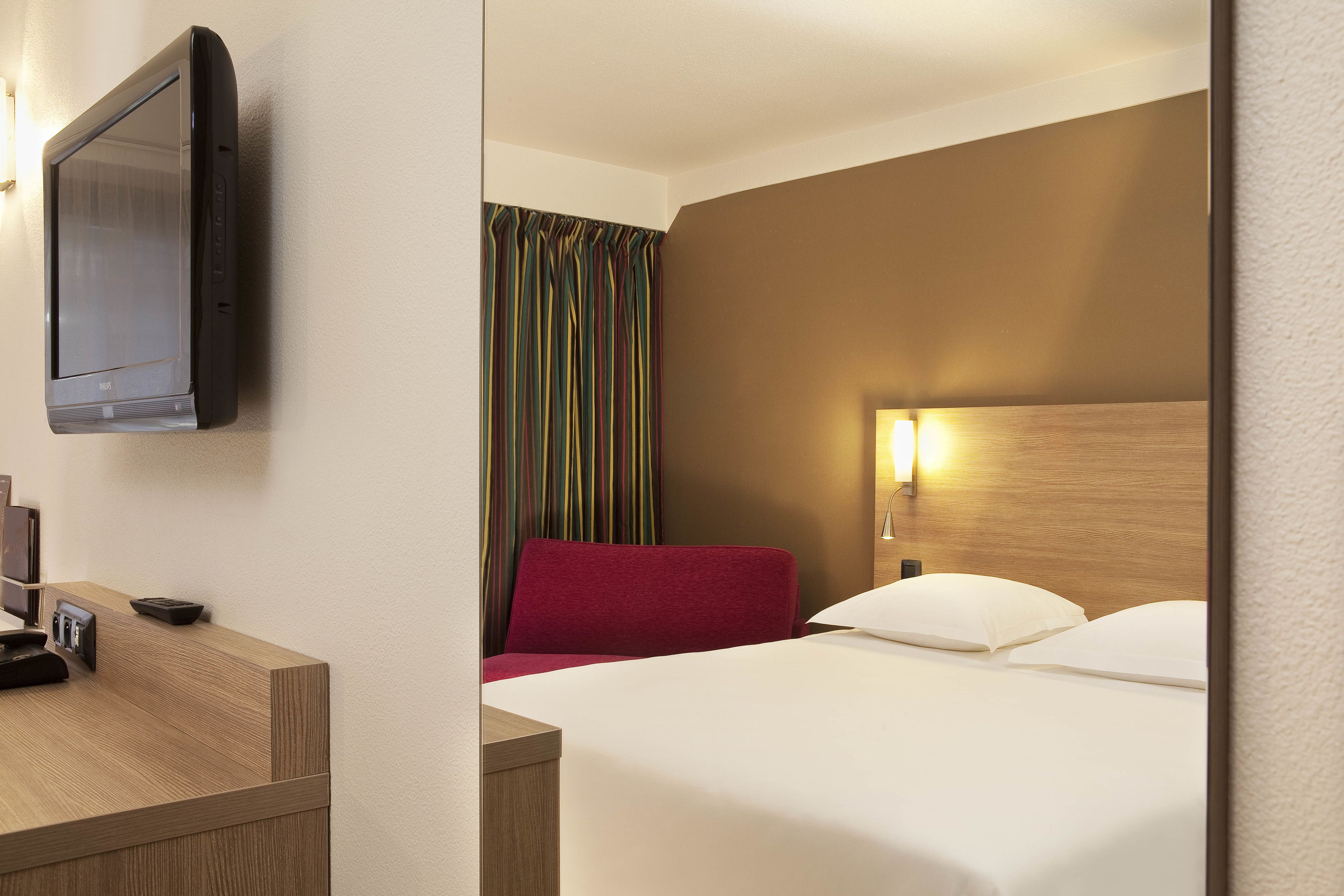Habitación de hotel con cama, escritorio, televisor de pantalla plana, una silla roja y cortina a rayas.