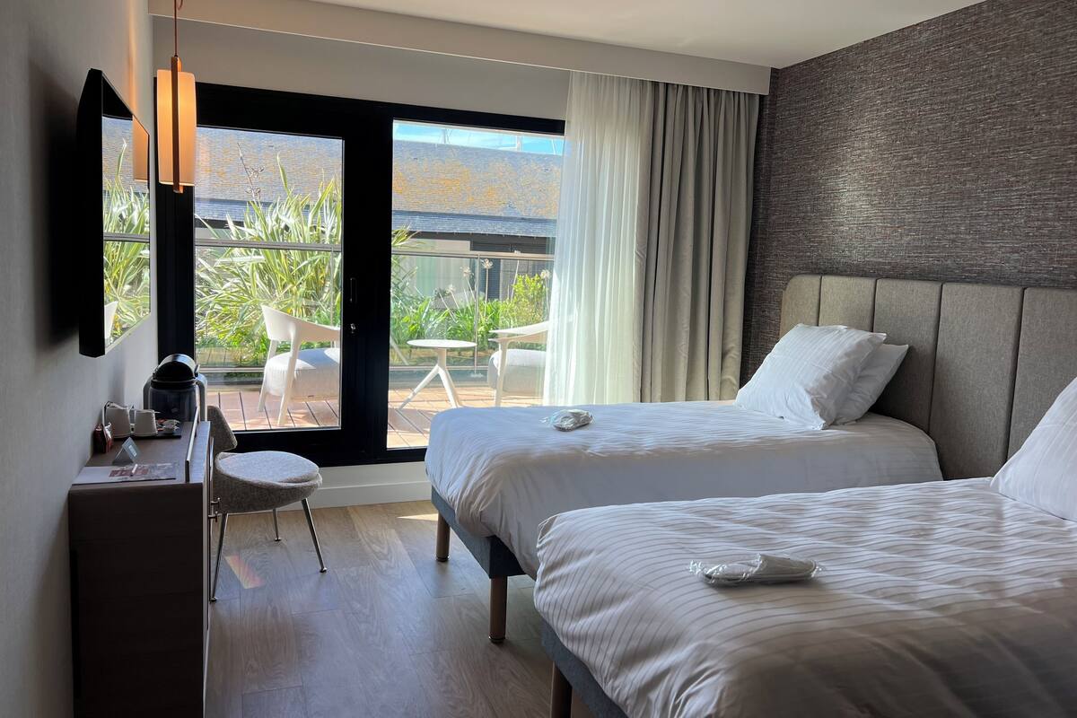 A hotel room with two beds, a desk, a chair, and a view of the outdoors.