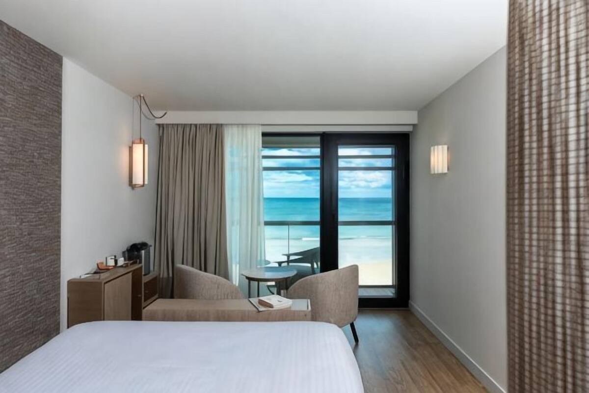 A hotel room with a bed, a desk, a chair, a small table, and a view of the ocean.