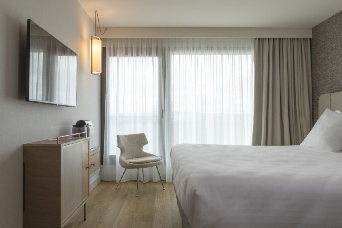 A hotel room with a bed, a chair, a dresser, a mirror, and a window with curtains.