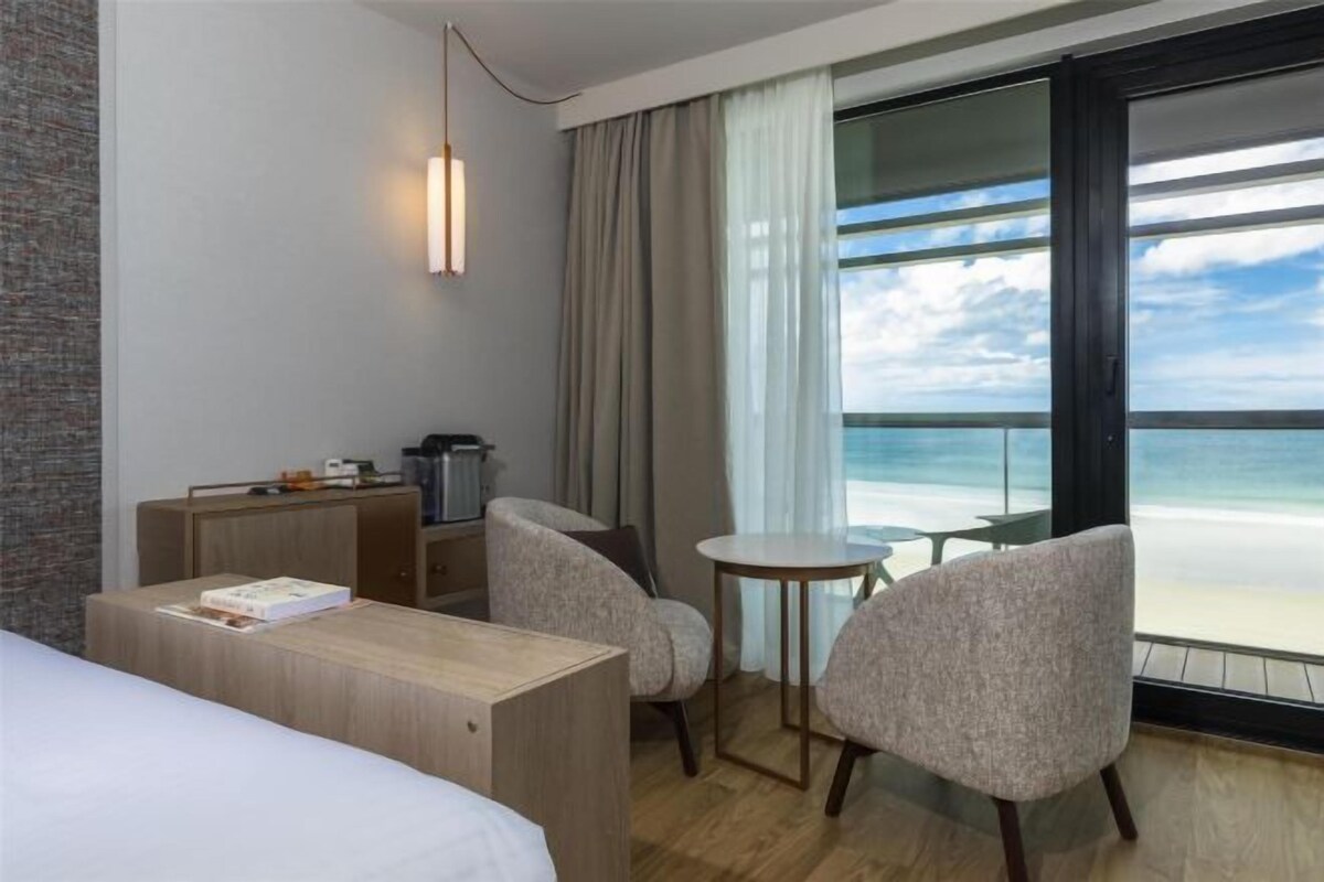 A hotel room with a bed, a desk, two chairs, a small table, and a large window offering a view of the beach.