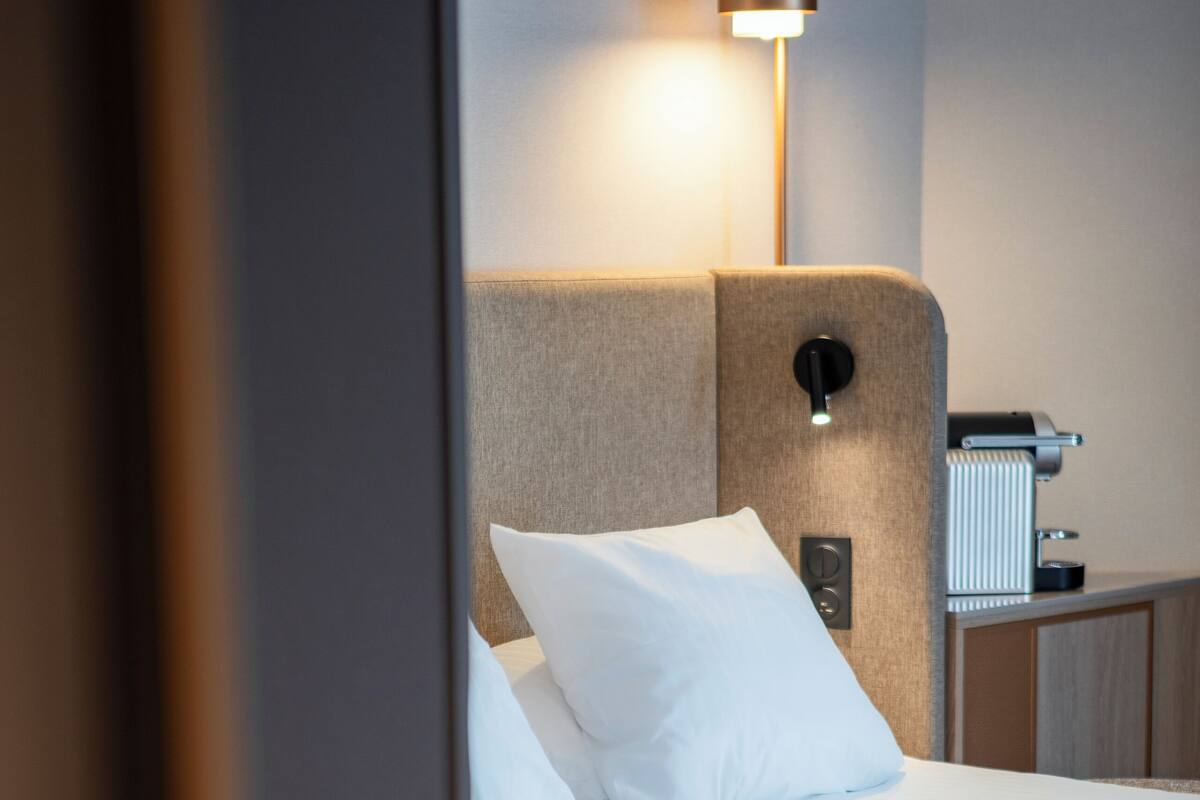 A neatly made bed with white linens, a headboard with a light switch, and a bedside table with a lamp.