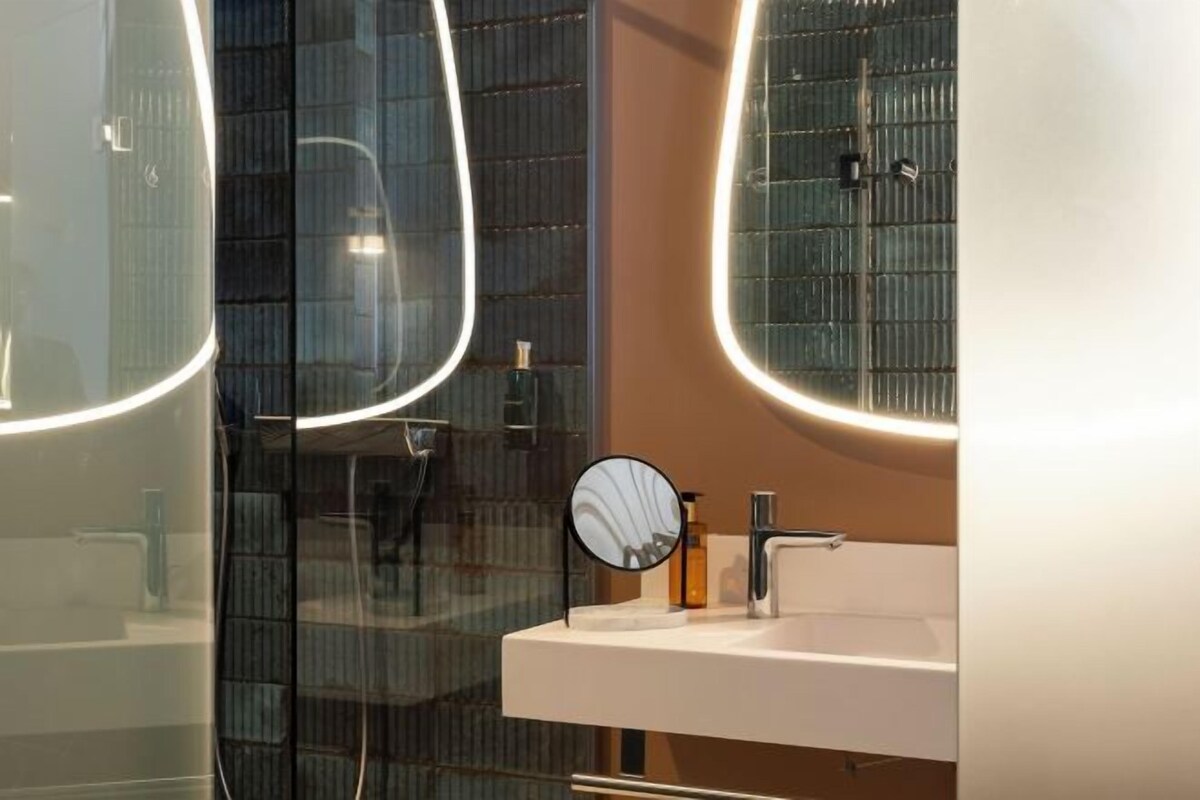 Un baño moderno con cabina de ducha de vidrio, un lavamanos blanco con espejo, y un espejo redondo en la pared.