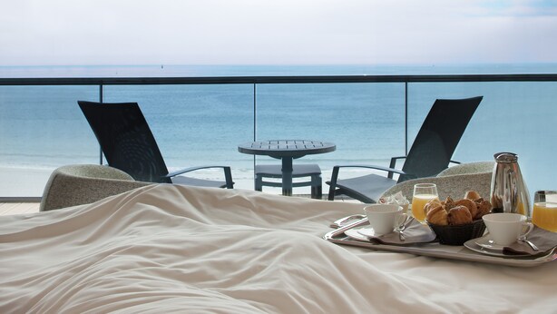 A balcony with a bed, a table with breakfast items, and a view of the sea. - Hotel Oceania Saint Malo (Saint-Malo)