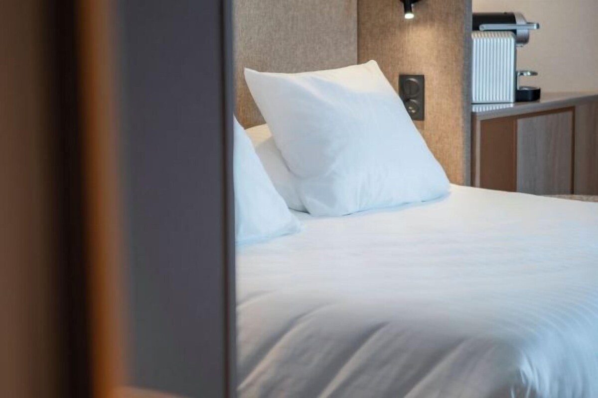 Una habitación de hotel con cama, almohadas, cabecera, mesita de noche y lámpara.
