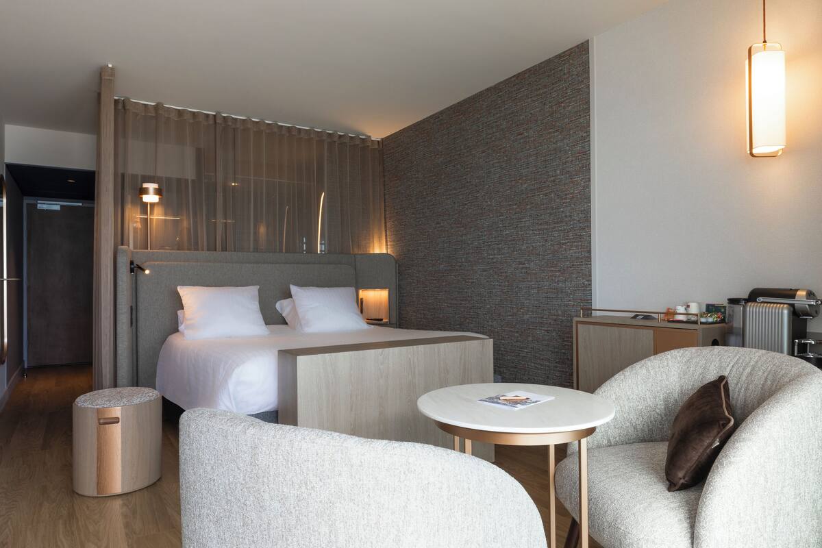 A modern hotel room with a bed, a small table, two armchairs, and a side table with a lamp.