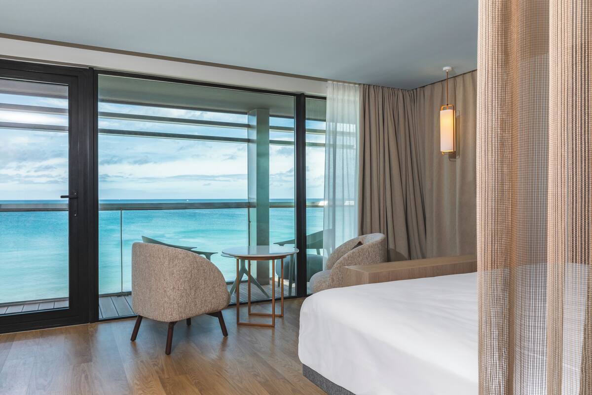 A hotel room with a large window, a bed, two armchairs, a small table, and a view of the ocean.