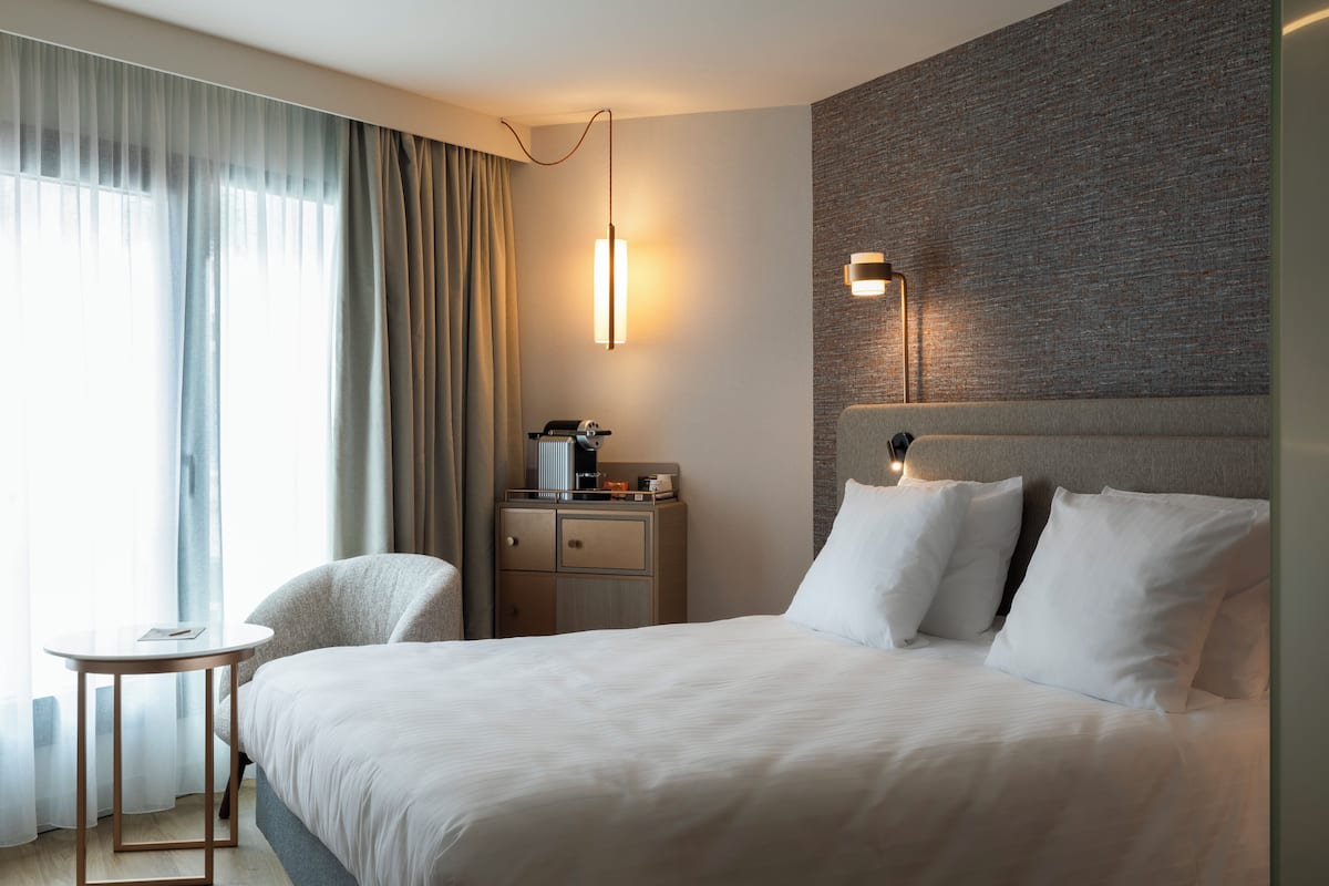A hotel room with a large bed, a bedside table, a chair, a desk, and a lamp.