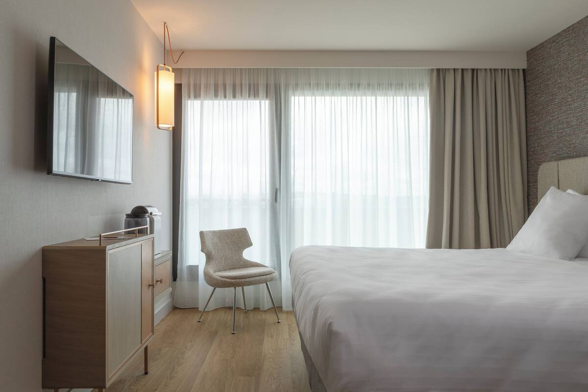 A modern hotel room with a large bed, a desk with a lamp, a chair, and a large window with curtains.