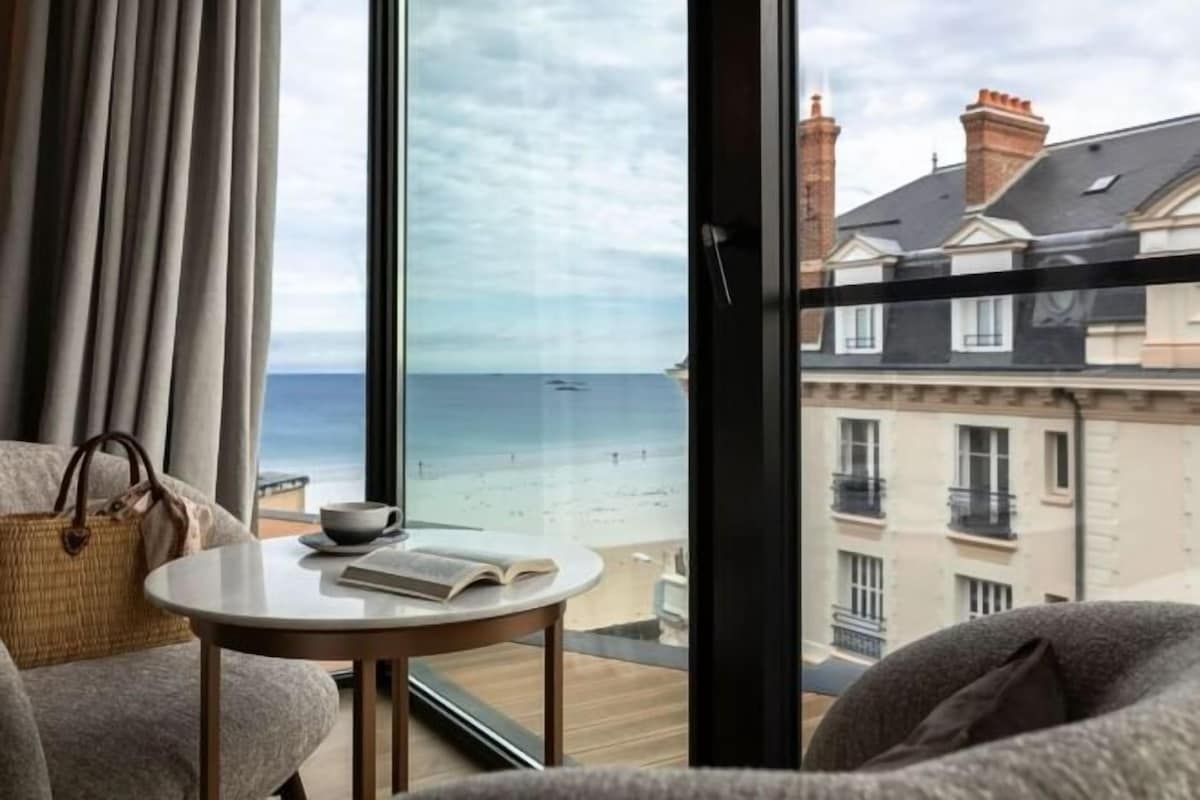 A room with a view of the beach, a round table with a book, and a window with curtains.