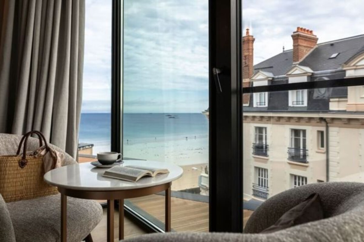 Quarto com vista para a praia, uma mesa redonda com um livro e uma janela com cortinas.