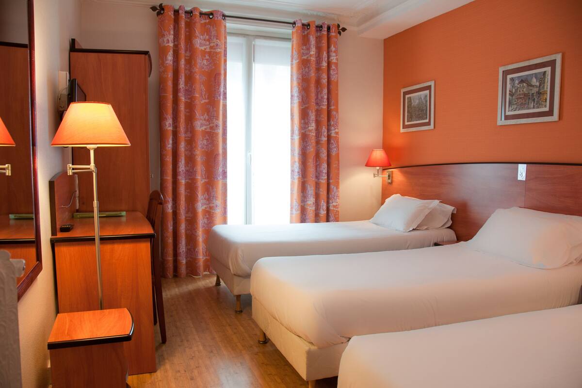 A hotel room with two single beds, a desk with a lamp, a chair, and a window with orange curtains.