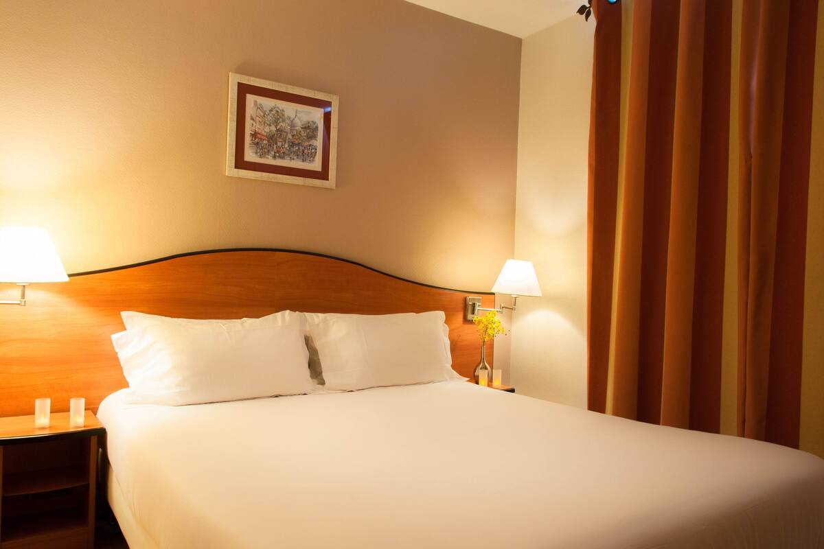 A hotel room with a large bed, two bedside lamps, a framed picture on the wall, and a curtain on the window.