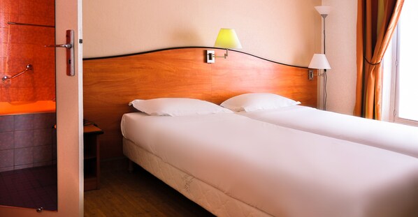 Une chambre d’hôtel avec une tête de lit en bois, un lit recouvert de draps blancs, deux lampes de chevet et une table de chevet.