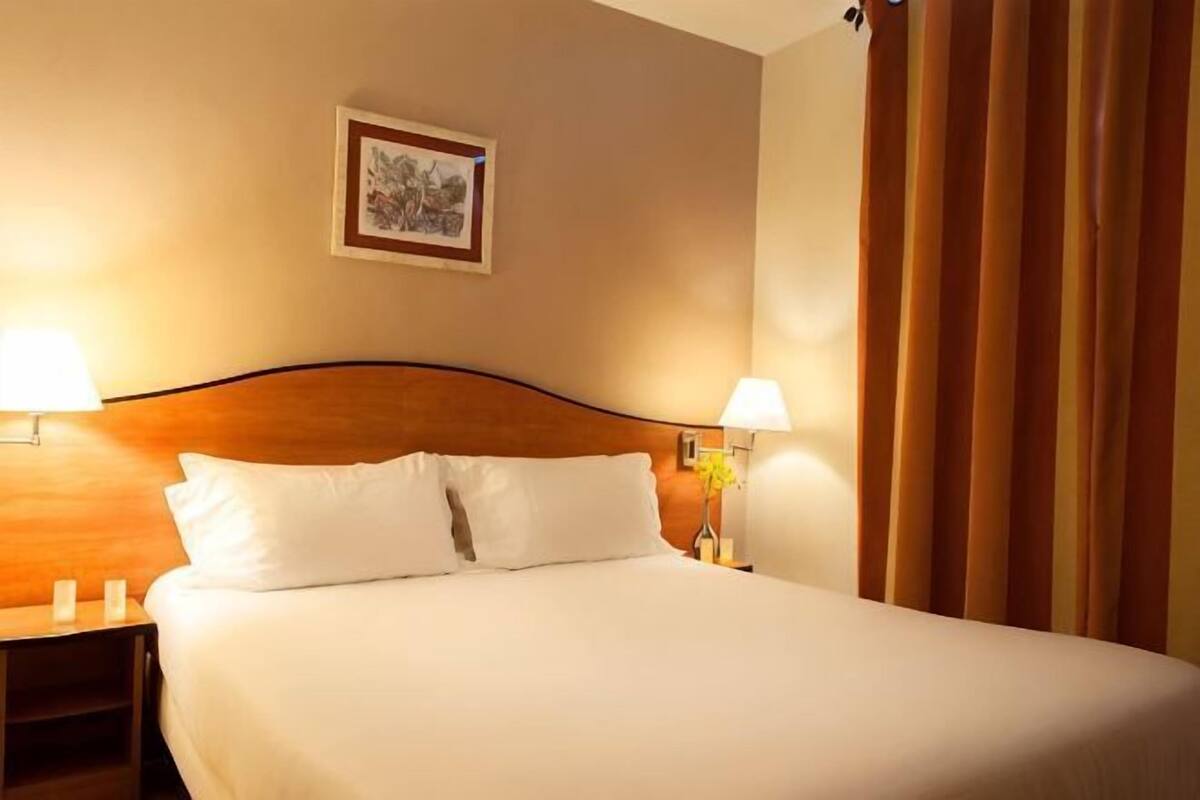 A neatly made bed with white linens, two bedside lamps, a wooden headboard, and a framed picture on the wall.