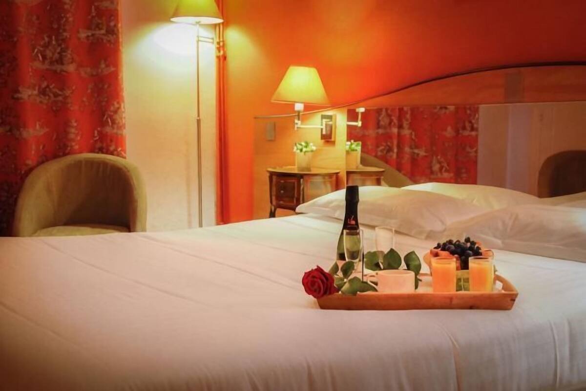 A hotel room with a bed, a bedside table, a lamp, and a tray with a bottle, a glass, and fruits.