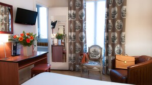 Een hotelkamer met een bed, een bureau met een stoel, een aan de muur bevestigde televisie, een raam met gordijnen en een stoel met een handtas.