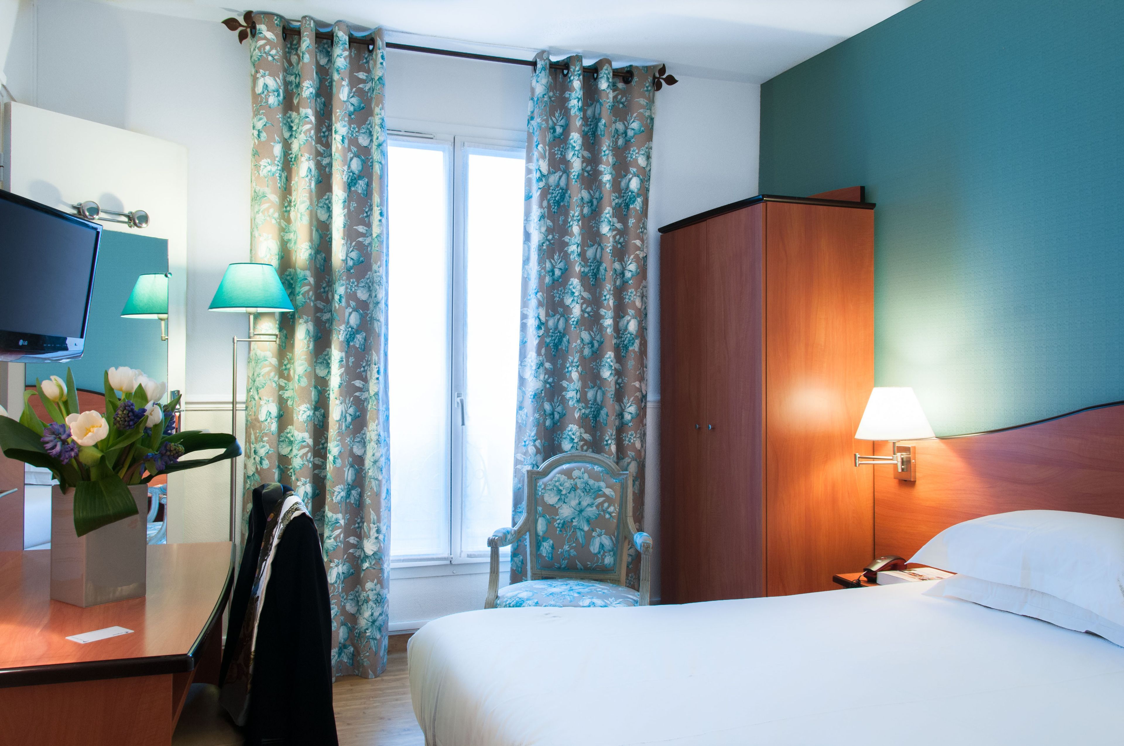 A hotel room with a bed, a desk with a TV, a chair, a wardrobe, and curtains.