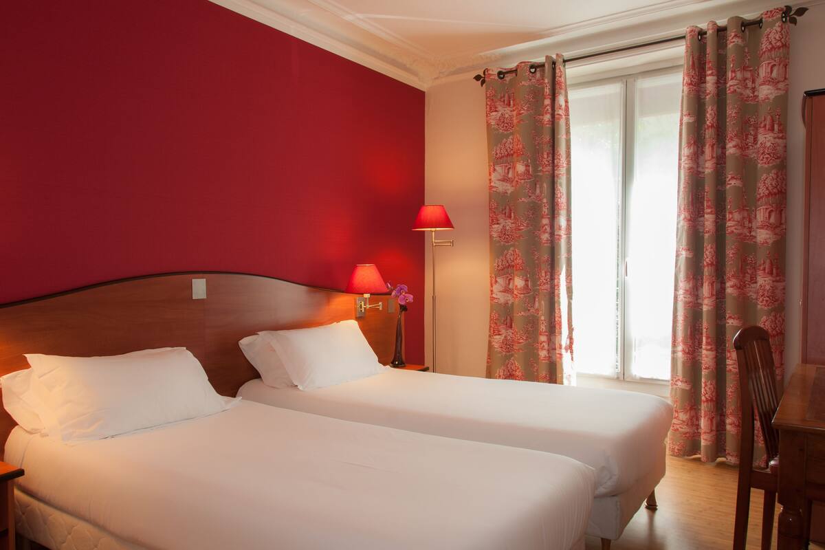 A hotel room with two beds, red walls, a window with floral curtains, and a wooden chair.