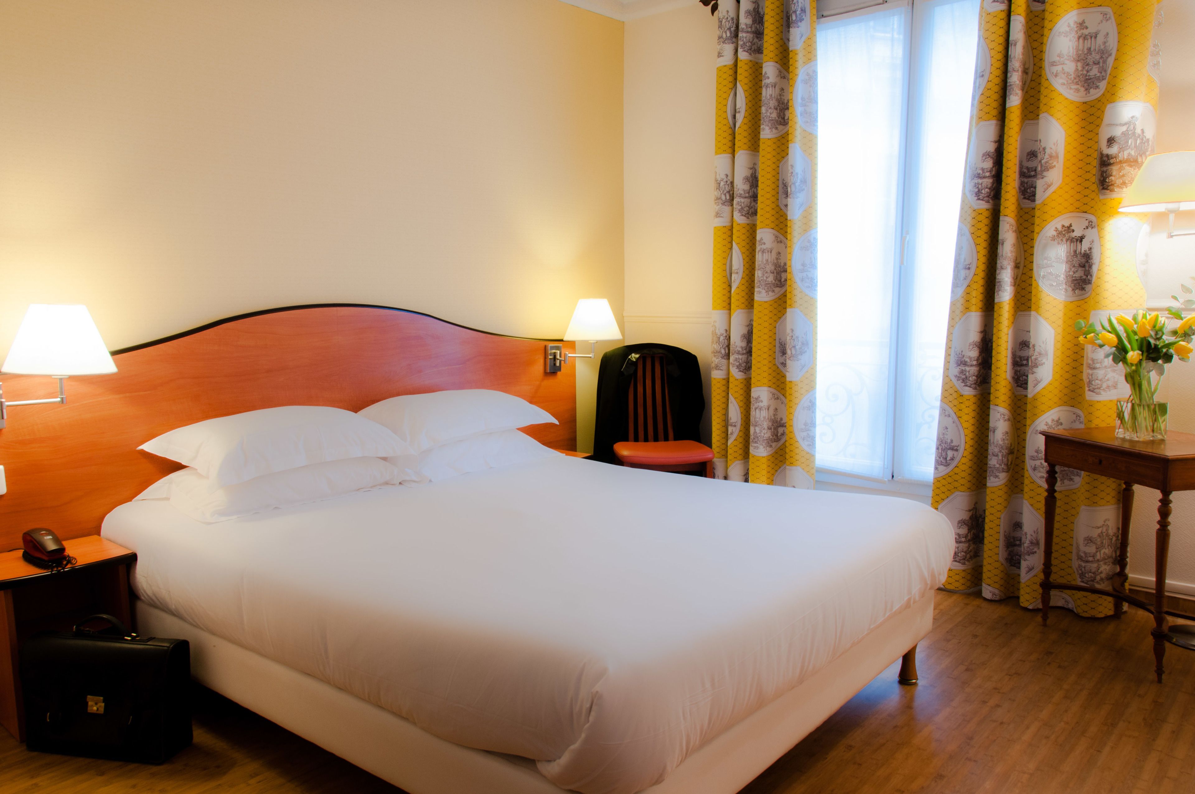 Una cama bien hecha con sábanas blancas, dos lámparas de noche, una mesita de noche con un teléfono y una maleta, y una ventana con cortinas amarillas.