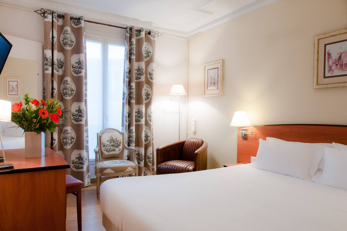 A hotel room with a bed, a desk with a lamp, a chair, and a vase with flowers.
