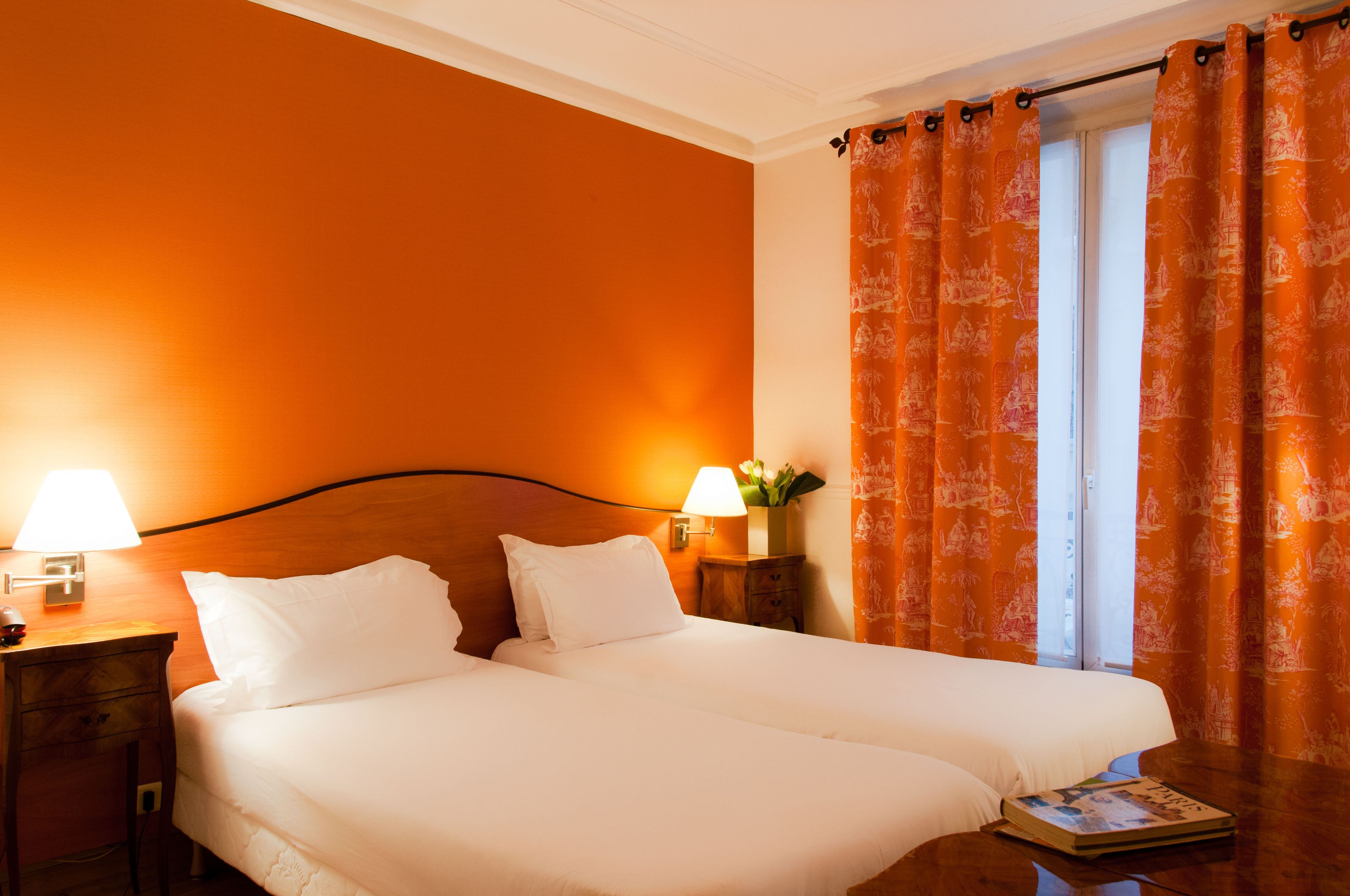 Una cama doble con sábanas blancas, dos lámparas de noche, una mesita de noche y una ventana con cortinas naranjas.