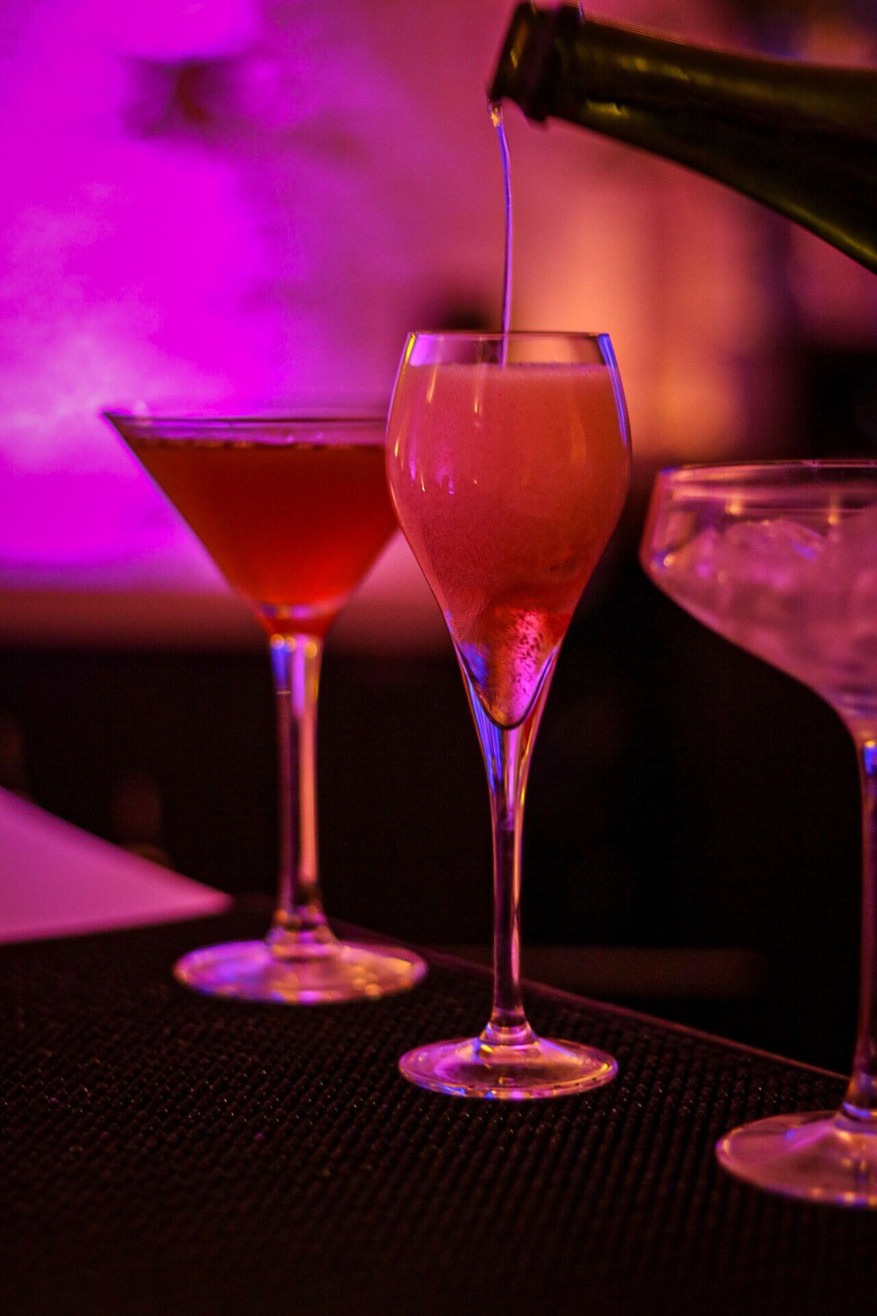 Three glasses of a pinkish beverage on a table with a textured surface, under purple lighting.