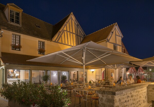 An outdoor dining area with tables and umbrellas, a stone wall, and a building with traditional architecture.