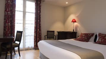 A hotel room with a bed, a desk, a chair, a lamp, and a view through a window with curtains.