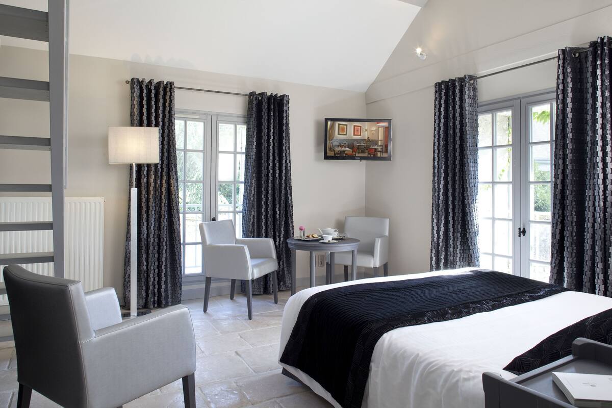 A hotel room with a bed, two chairs, a small table with a cup and saucer, and curtains.
