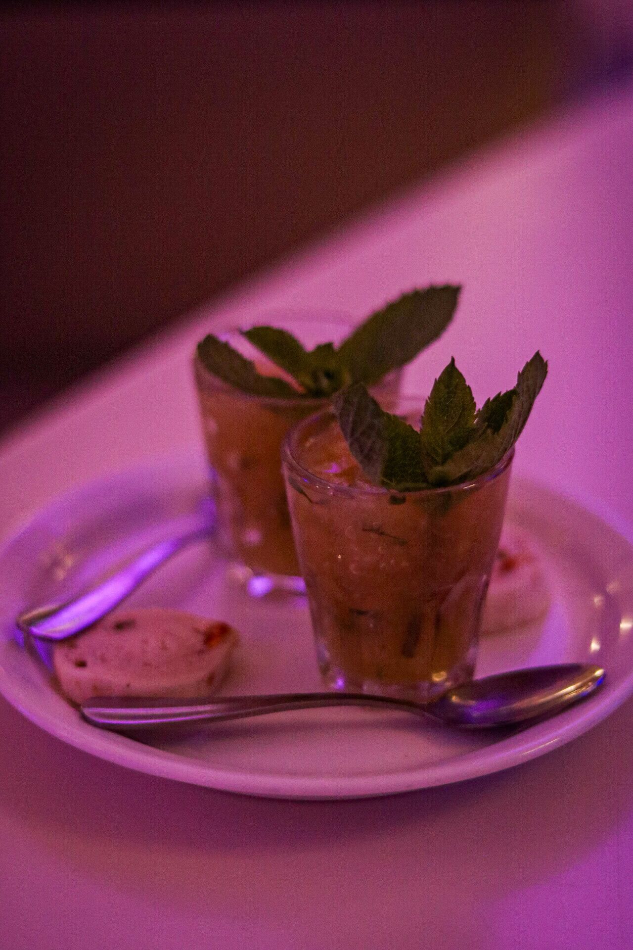 Two glasses of a beverage with mint leaves on a white plate, accompanied by a spoon.