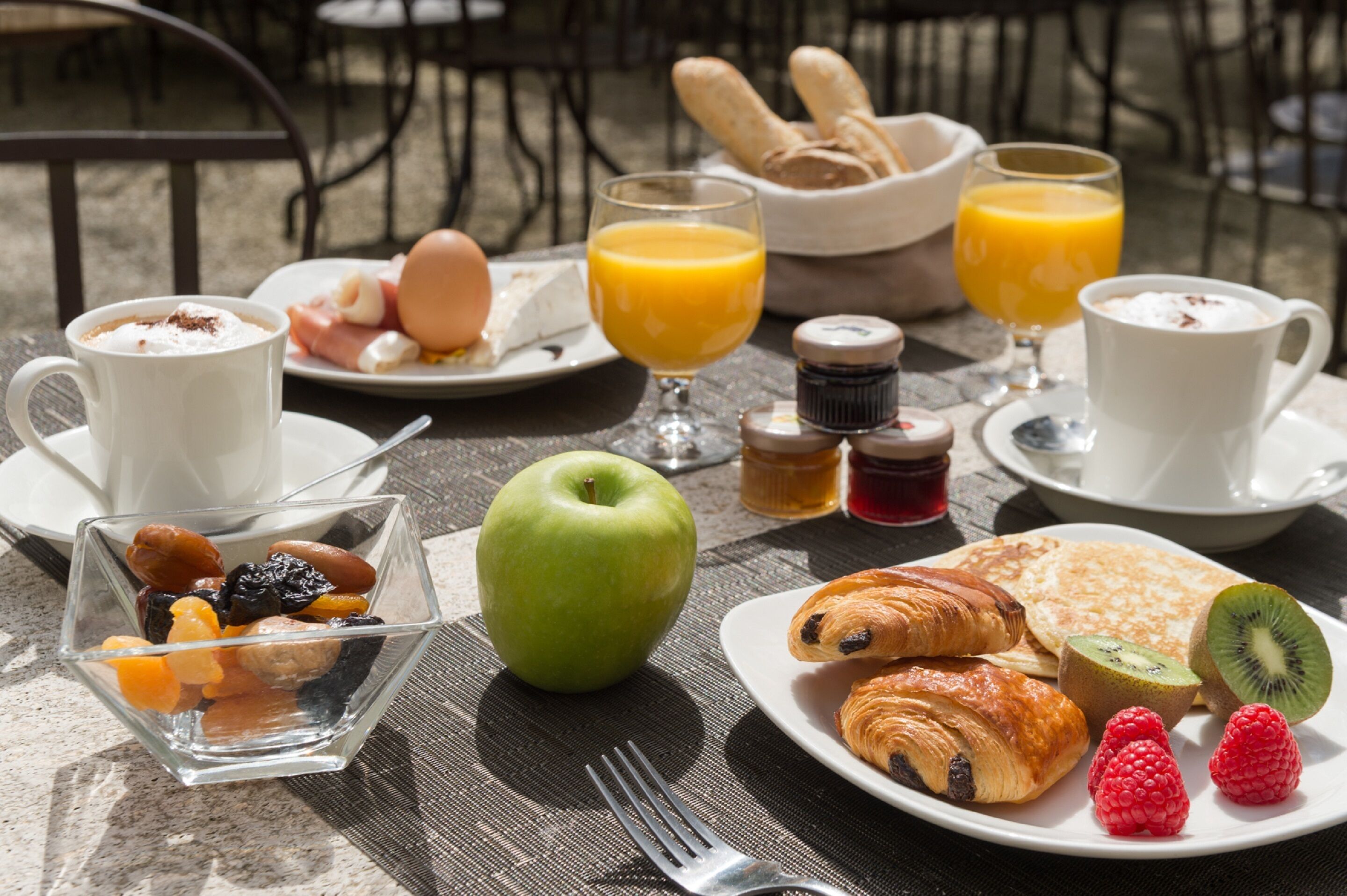 Une table de petit-déjeuner avec des croissants, des fruits, un verre de jus d’orange, une tasse de café et une assiette d’œufs et de pain.