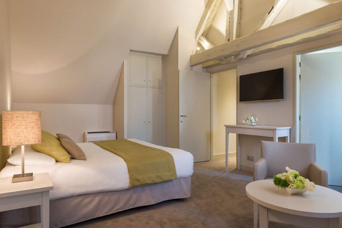 Quarto de hotel com cama, mesa de cabeceira, luminária, cadeira, escrivaninha, televisão e uma pequena mesa com flores.