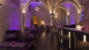 Un interior tenuemente iluminado, con paredes y arcos de piedra, que presenta una zona de bar con taburetes y una botella de vino.