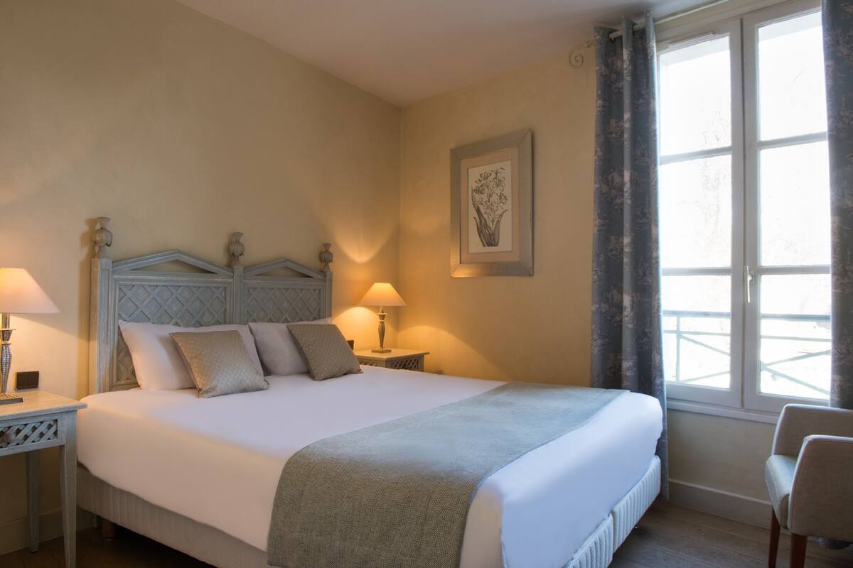 A neatly made bed with a headboard, two bedside tables with lamps, a framed artwork on the wall, and a window with curtains.