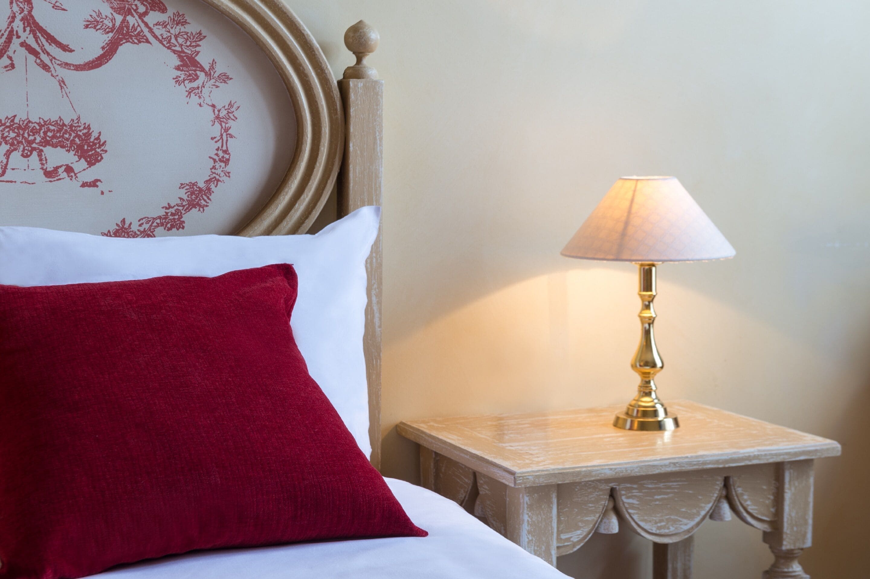 A neatly made bed with a red pillow and a white pillow, a small bedside table with a lamp, and a wall with a floral design.