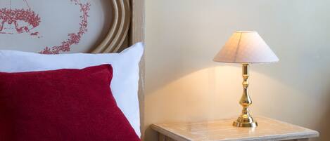 A neatly made bed with a red pillow and a white pillow, a small bedside table with a lamp, and a wall with a floral design.