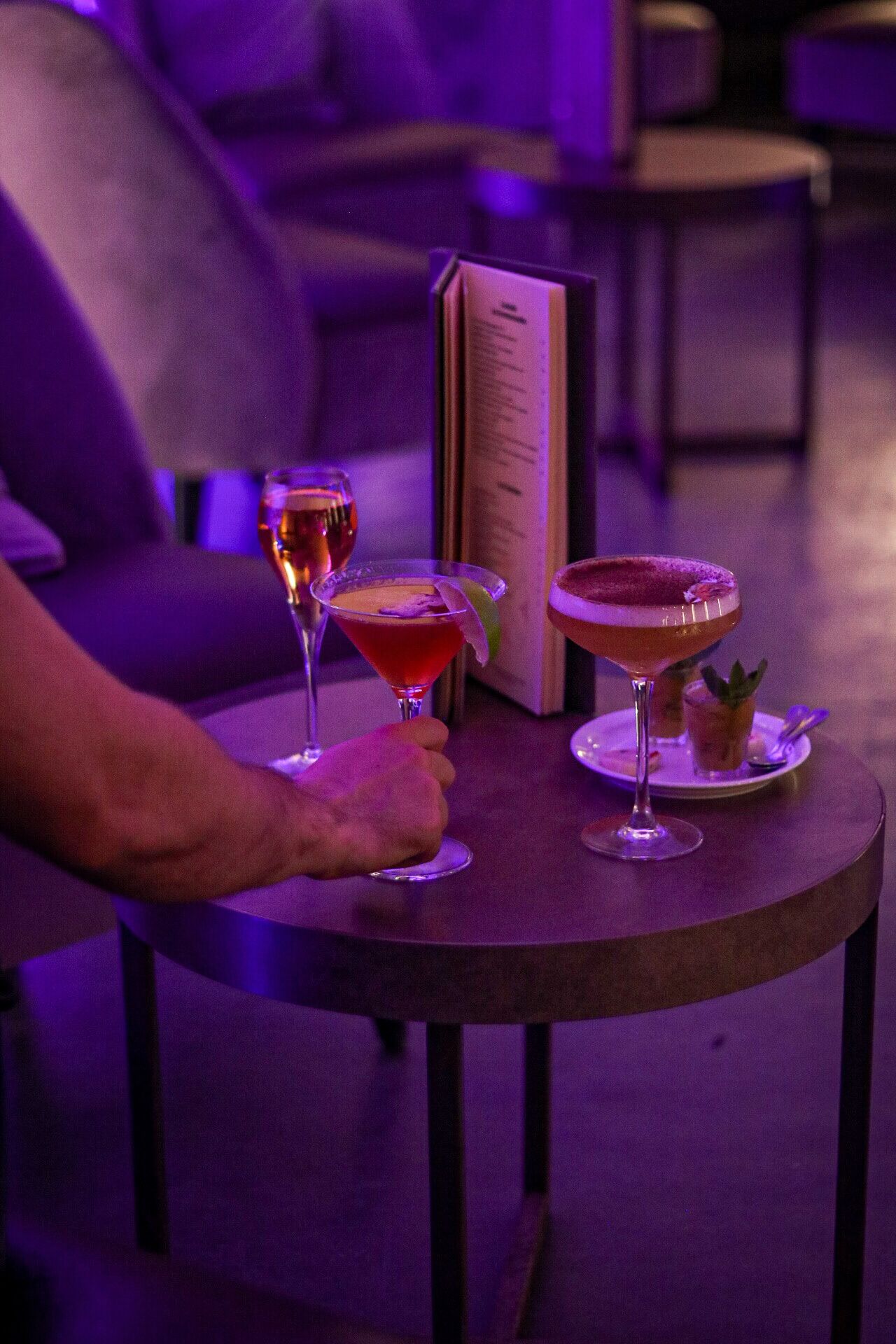 A table with three drinks, a menu, and a person sitting on a couch.