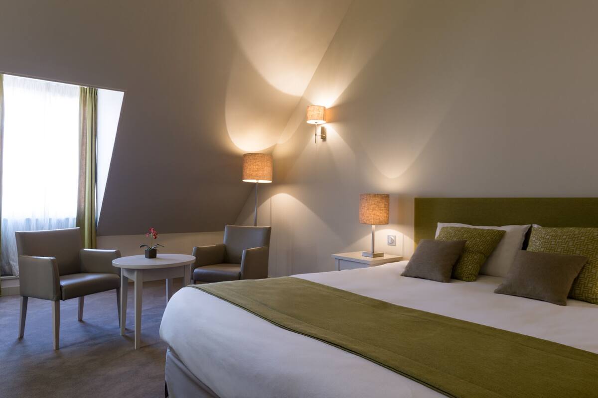 A hotel room with a bed, two chairs, a small table, and two wall-mounted lamps.