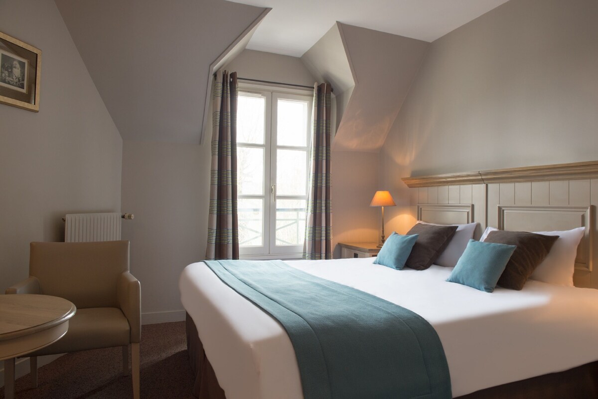 Quarto de hotel com uma cama grande, uma mesa redonda, uma cadeira, uma janela com cortinas e um quadro na parede.
