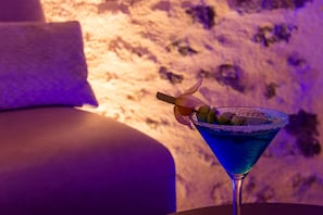 A cocktail in a martini glass with a salted rim, garnished with olives and a cherry, on a table with a purple-lit background.