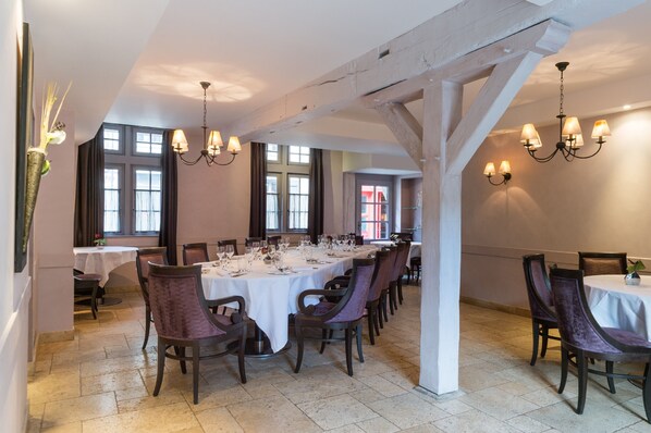 A dining area with multiple tables set for a meal, featuring chandeliers and a view of the outside through large windows.