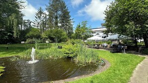 A park with a pond, outdoor seating, and a building with 'VELTINS' signage. - VitalHotel Ascona (Bad Bevensen)