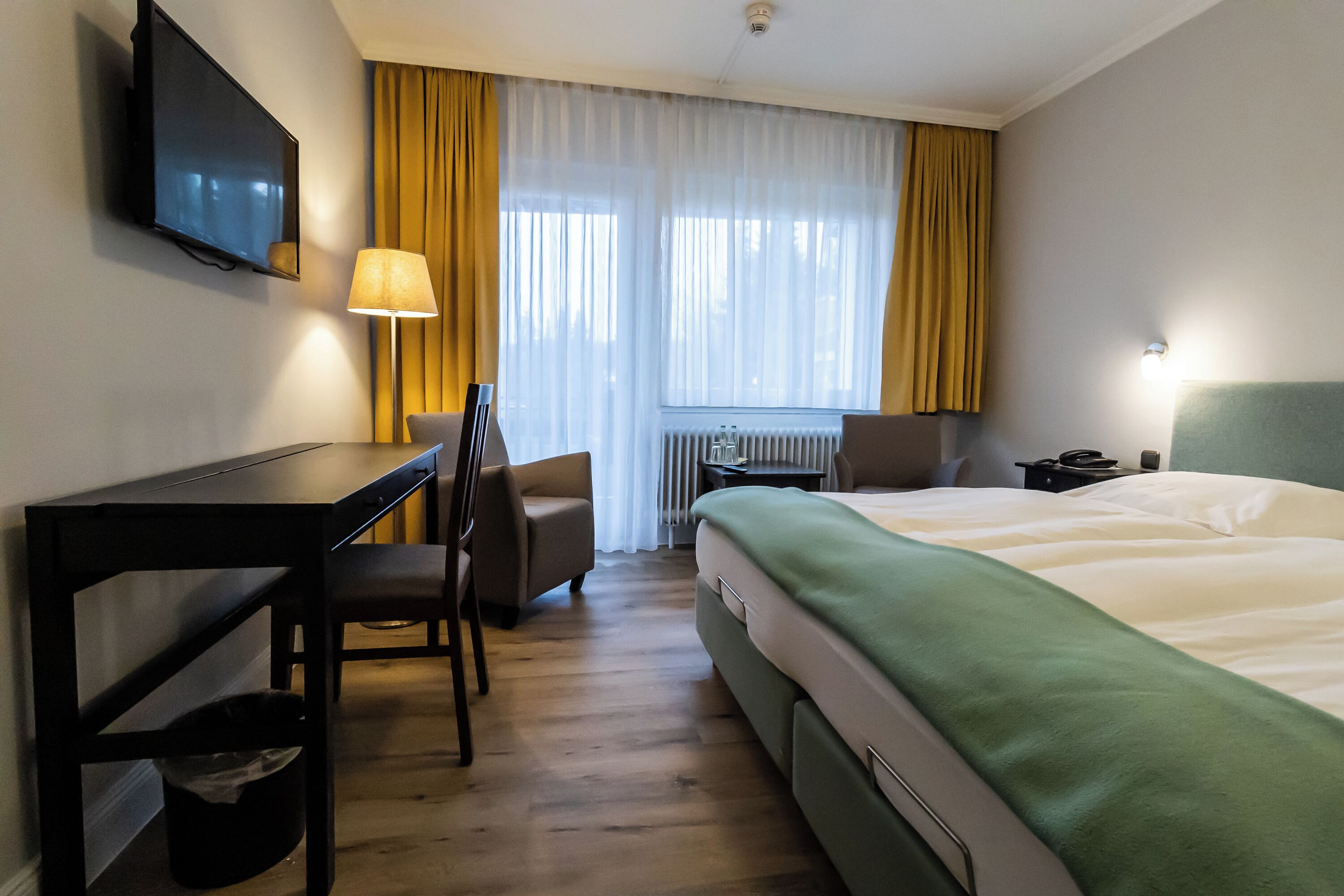 Habitación de hotel con cama, escritorio, silla, televisor y ventana con cortinas.
