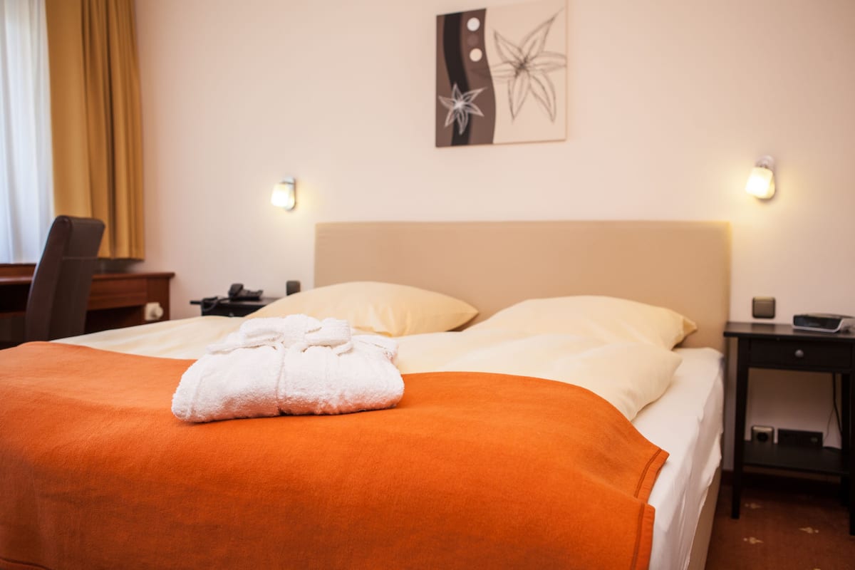 A neatly made bed with an orange blanket and a white towel, a bedside table with a lamp, and a wall art piece.