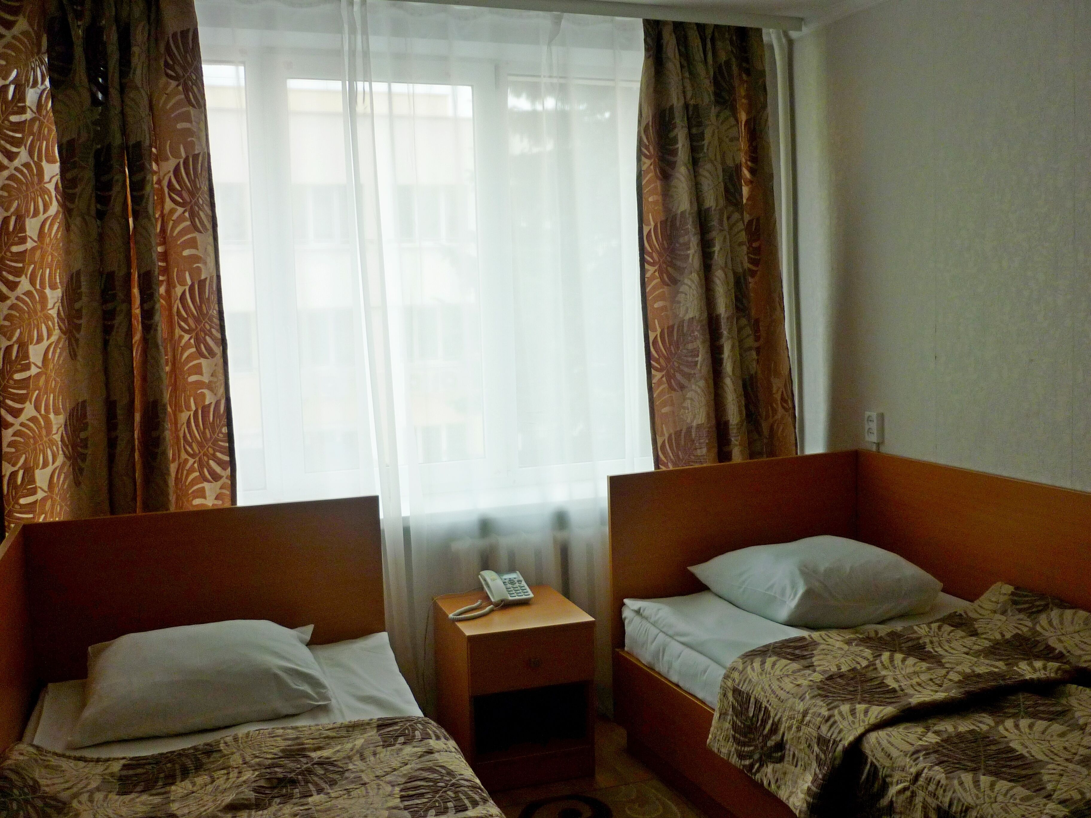 A hotel room with two single beds, a bedside table with a phone, and a window with sheer curtains.